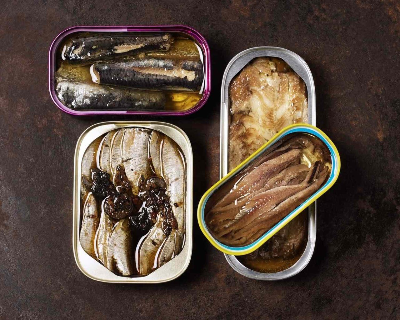 Assorted tinned seafood, including sardines, anchovies, and other fish in oil, displayed in open cans.