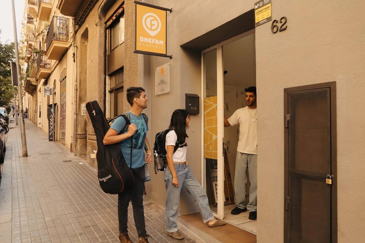 Two travelers entering the Onefam Paralelo Hostel in Barcelona, greeted by a staff member at the entrance.