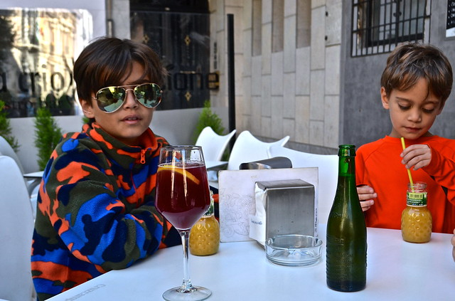 Two kids sitting at an outdoor café in Barcelona, enjoying drinks with one child wearing sunglasses and a colorful jacket.