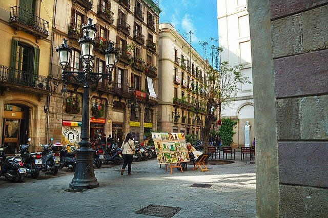 A quiet square in Barcelona with a local artist displaying paintings and scooters parked nearby.