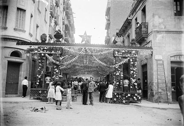 Black-and-white photo of a decorated street during a historical edition of the Festa Major de Gràcia in Barcelona, with people admiring the floral and festive ornaments.