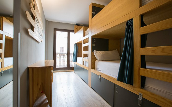 Modern hostel room with wooden bunk beds, privacy curtains, light wood floors, and a balcony view.