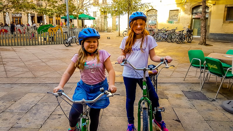 Two kids riding bikes with helmets in a plaza in Barcelona, enjoying a sunny day near an outdoor café.