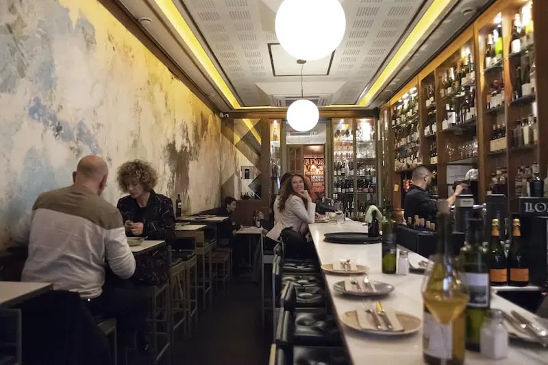Denassus wine bar in Barcelona with a modern, intimate interior, featuring a long bar counter, extensive wine displays, and patrons enjoying their evening.