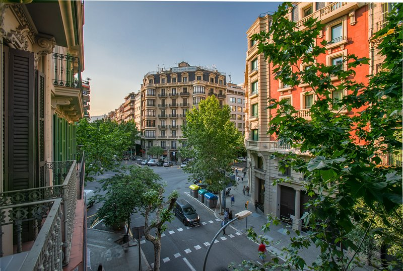 A view of a typical intersection in Barcelona's Eixample neighborhood, showcasing the area's distinctive grid layout, ornate architecture, and lush green trees.