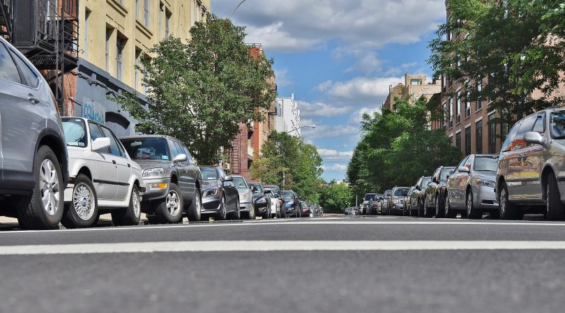 A city street lined with parked cars on both sides, viewed from ground level. Buildings with trees and a partly cloudy blue sky create a peaceful urban scene.